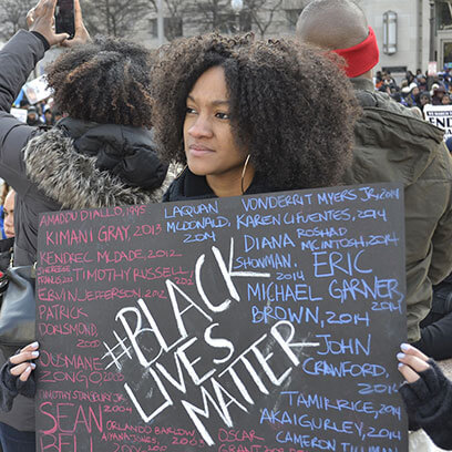 A protester holds a handmade sign at a Black Lives Matter demonstration. The sign features the large, bold hashtag '#BLACK LIVES MATTER' surrounded by the handwritten names of Black individuals killed by police or vigilantes, along with the years of their deaths. The crowd in the background adds to the powerful atmosphere of collective mourning and resistance.