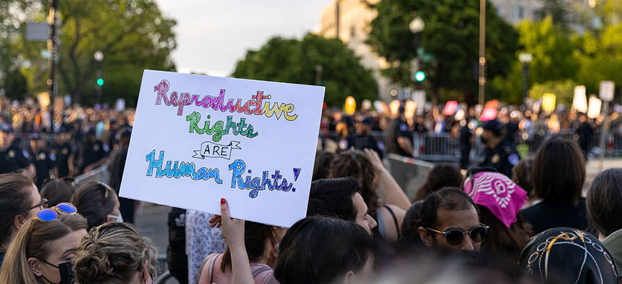 A protester holds a colorful handmade sign reading ‘Reproductive Rights ARE Human Rights!’ during a large outdoor demonstration. The crowd is gathered closely together, with police and barricades visible in the background, set against a backdrop of trees and city buildings.