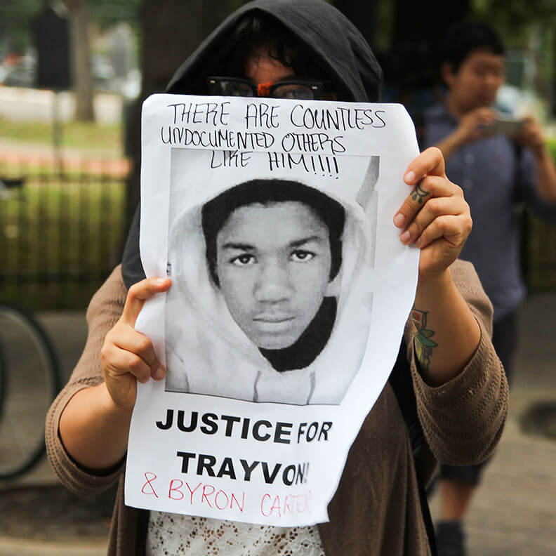 A person wearing a hoodie holds up a sign featuring a black-and-white photo of Trayvon Martin. The top of the sign reads, 'THERE ARE COUNTLESS UNDOCUMENTED OTHERS LIKE HIM!!!!' and the bottom reads, 'JUSTICE FOR TRAYVON!' with '& BYRON CARTER' handwritten underneath. The image captures a moment of protest and solidarity in the fight for racial justice.